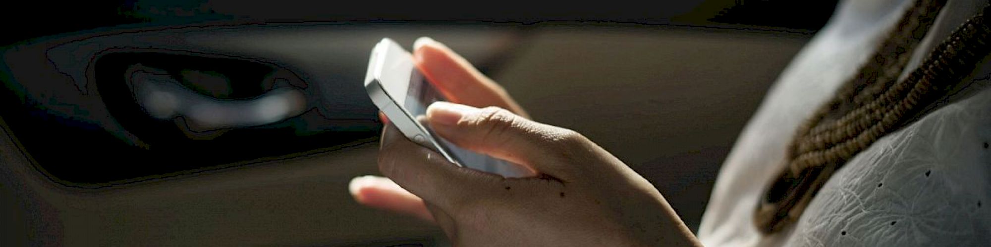 A person is using a smartphone while sitting in a vehicle. The focus is on the hands holding the phone, with a blurred car interior in the background.