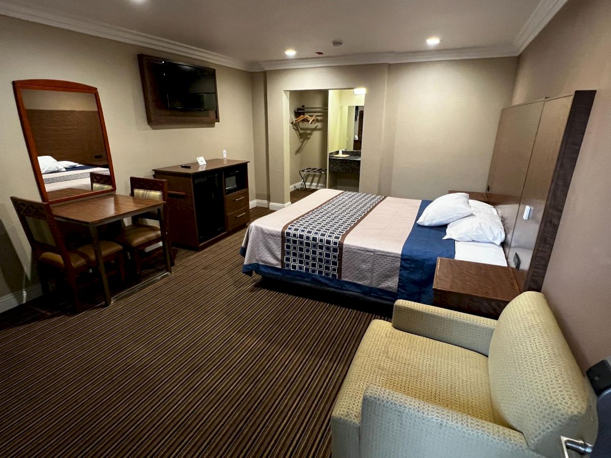 A hotel room with a bed, armchair, desk, and TV. There's also a mirror, mini fridge, and wardrobe in a cozy setting.