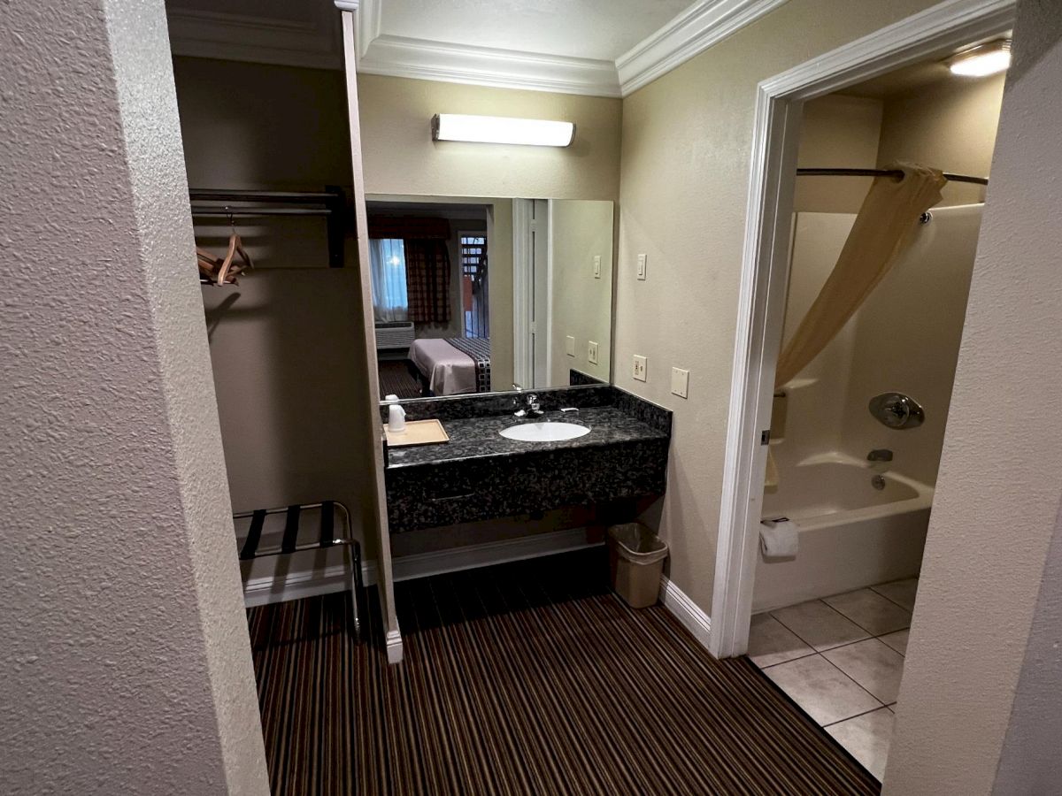 The image shows a hotel room bathroom with a sink, mirror, closet, toilet, and bathtub with a shower curtain in a compact space.