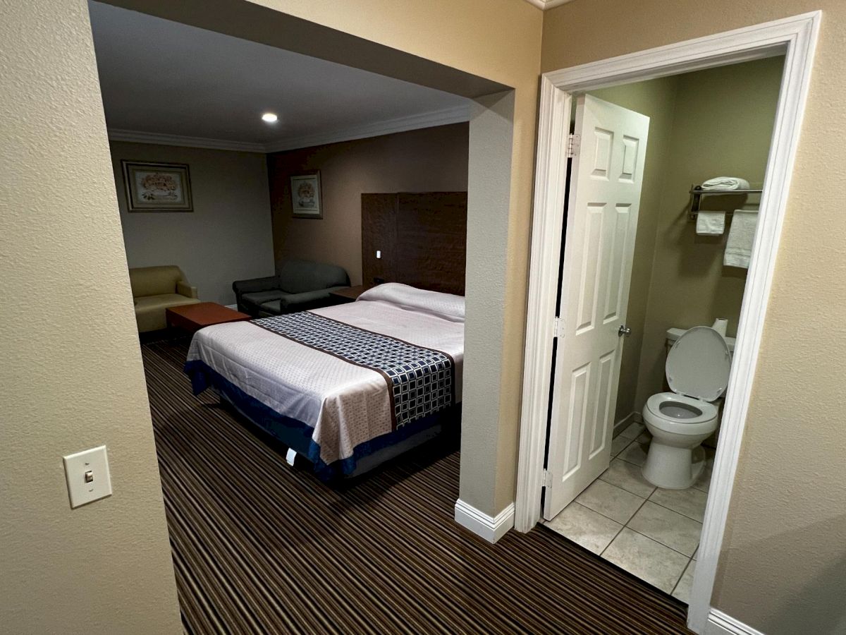 A hotel room with a bed, a small sitting area, and an open door to a bathroom with a toilet and towel rack visible.