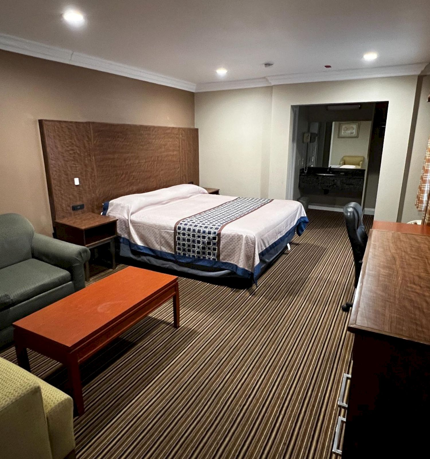 This image shows a hotel room with a bed, sofa, coffee table, desk, TV, and artwork on the wall, featuring striped carpet flooring.