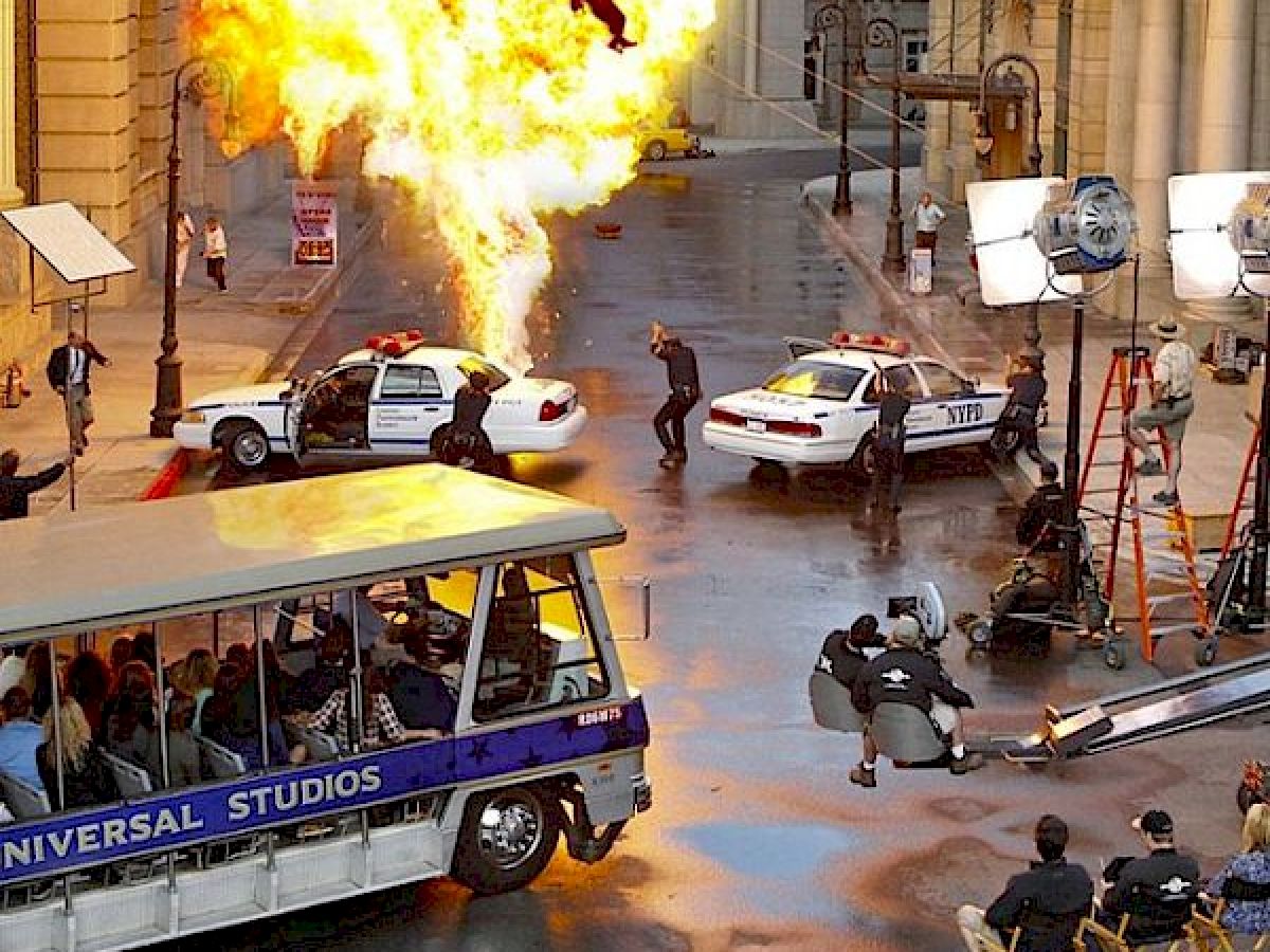 This image shows a Universal Studios tour tram passing a movie set, featuring an explosion scene with police cars, actors, and film crew members.