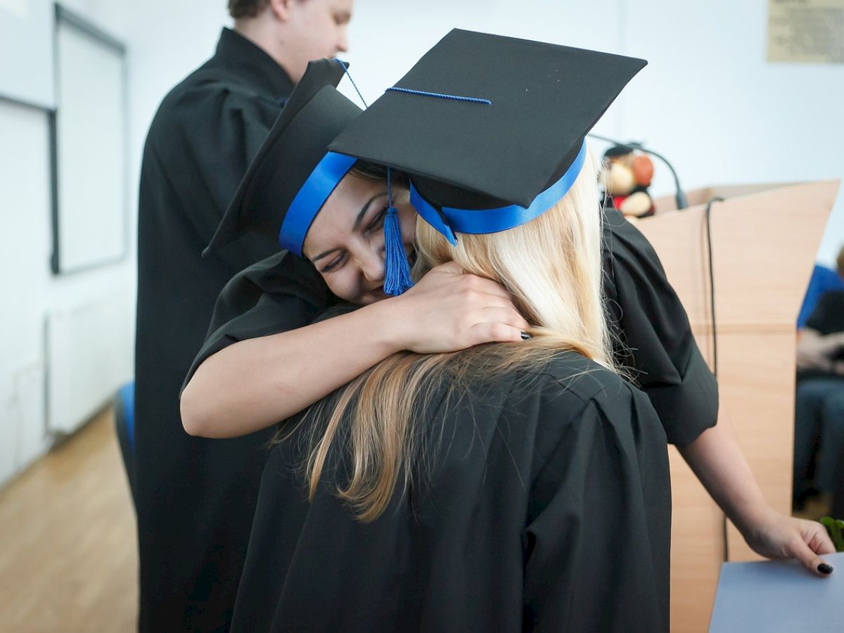 Two graduates in caps and gowns are hugging each other, with one smiling widely, while another person stands nearby in the background.