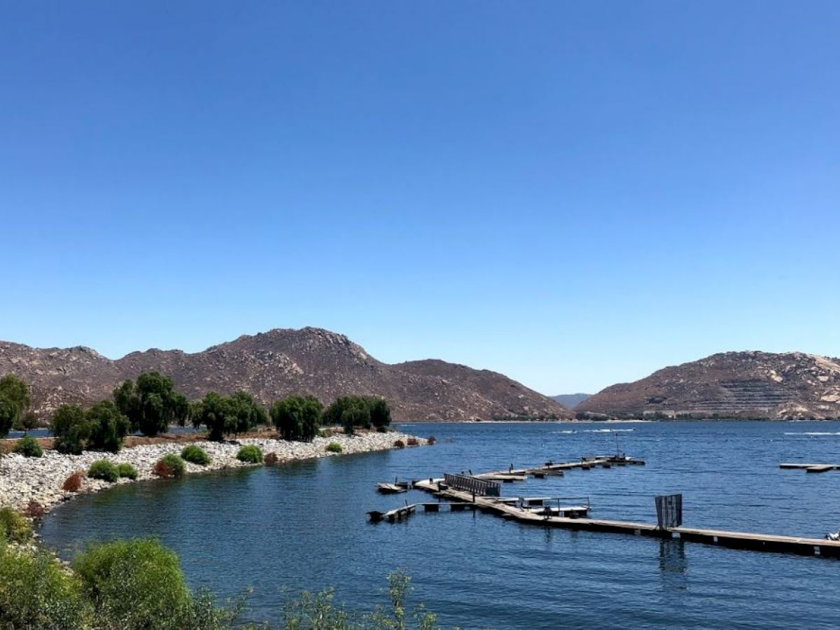 A serene lake with clear blue water, surrounded by hills and greenery, features a few docks extending into the water under a clear blue sky.