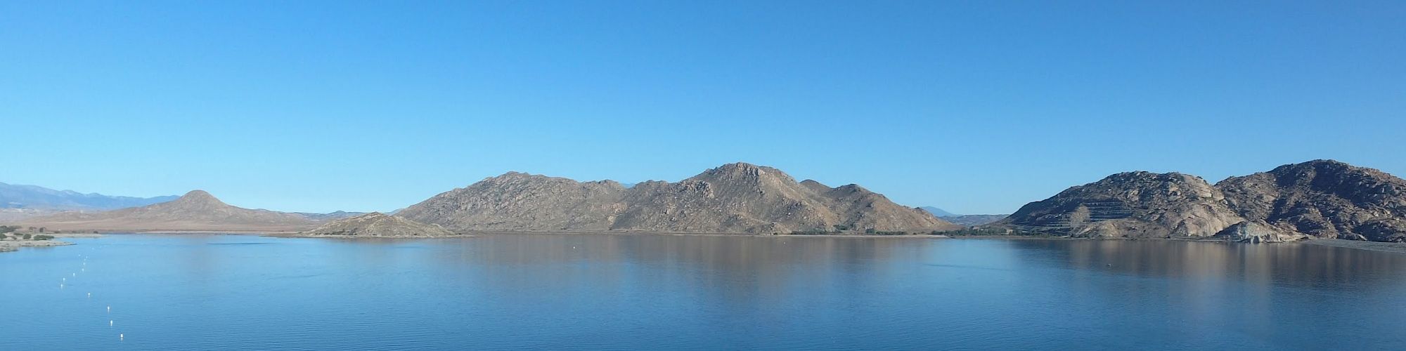 A serene lake with a rocky shore and distant mountains under a clear blue sky.