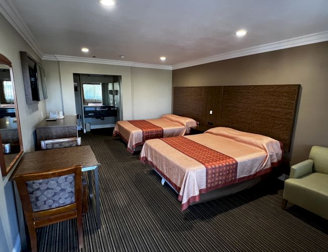 The image shows a hotel room with two beds, a mirror, desk, chair, TV, and an armchair, featuring neutral tones and modern decor.