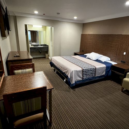 This image shows a hotel room with a double bed, desk, chair, and side tables. The room is well-lit with simple, modern furnishings.