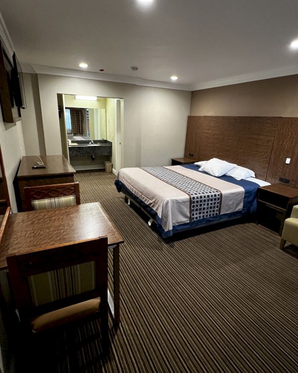 This image shows a hotel room with a double bed, desk, chair, and side tables. The room is well-lit with simple, modern furnishings.