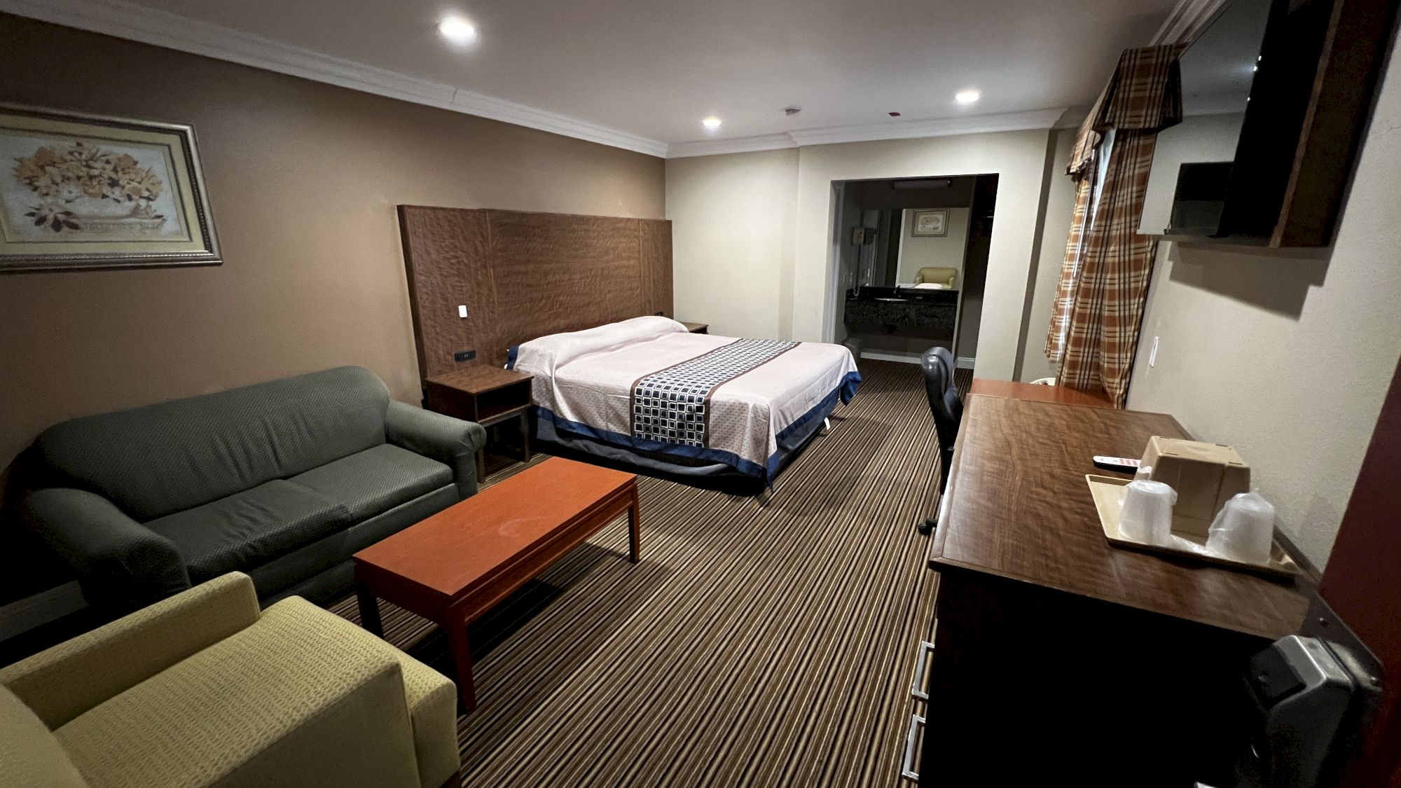 The image shows a hotel room with a bed, two sofas, a desk, TV, and nightstand, decorated in neutral tones with striped carpet flooring.