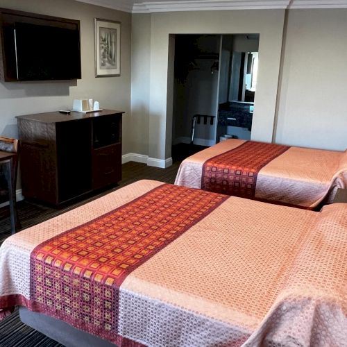 The image shows a hotel room with two beds, a TV, a small table with chairs, and a dresser. The room has a mirror and simple decor.