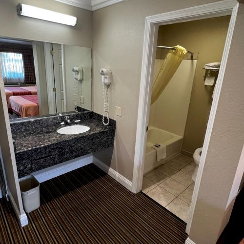 The image shows a hotel bathroom with a sink, mirror, and hairdryer. There's a separate area with a bathtub, toilet, and towels.