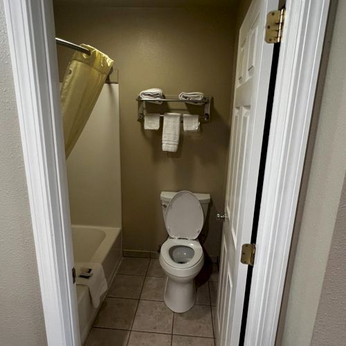 The image shows a small bathroom with a toilet, bath with shower curtain, and towels neatly stacked on a rack above the toilet.