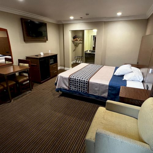 The image shows a hotel room with a bed, armchair, table, chairs, dresser, flatscreen TV, and a mirror. Neutral-colored decor is visible.