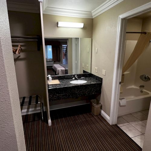 The image shows a hotel room bathroom area with a sink, mirror, open closet, and a shower-tub with a curtain.