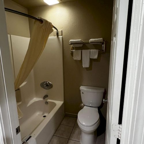 The image shows a small bathroom with a bathtub, shower curtain, toilet, and wall-mounted towels. The floor is tiled, and lighting is overhead.