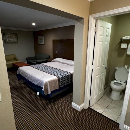 The image shows a hotel room with a neatly made bed, a sitting area, and an adjacent bathroom with a toilet visible.