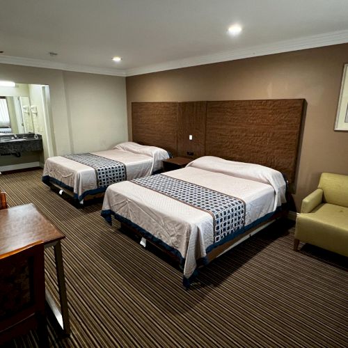 The image shows a hotel room with two double beds, a desk, a chair, and an air conditioner. The decor is neutral and cozy.