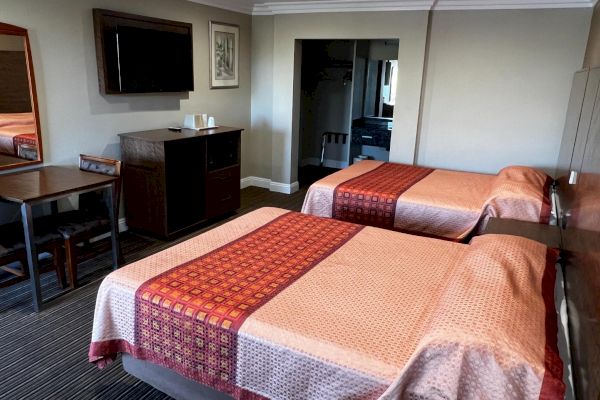 The image shows a hotel room with two beds, a TV, a small table with chairs, and a mirror on the wall. The decor is simple and neat.