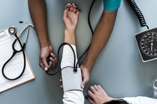 A healthcare professional is measuring a patient's blood pressure with a sphygmomanometer on a desk, with a stethoscope and clipboard nearby.